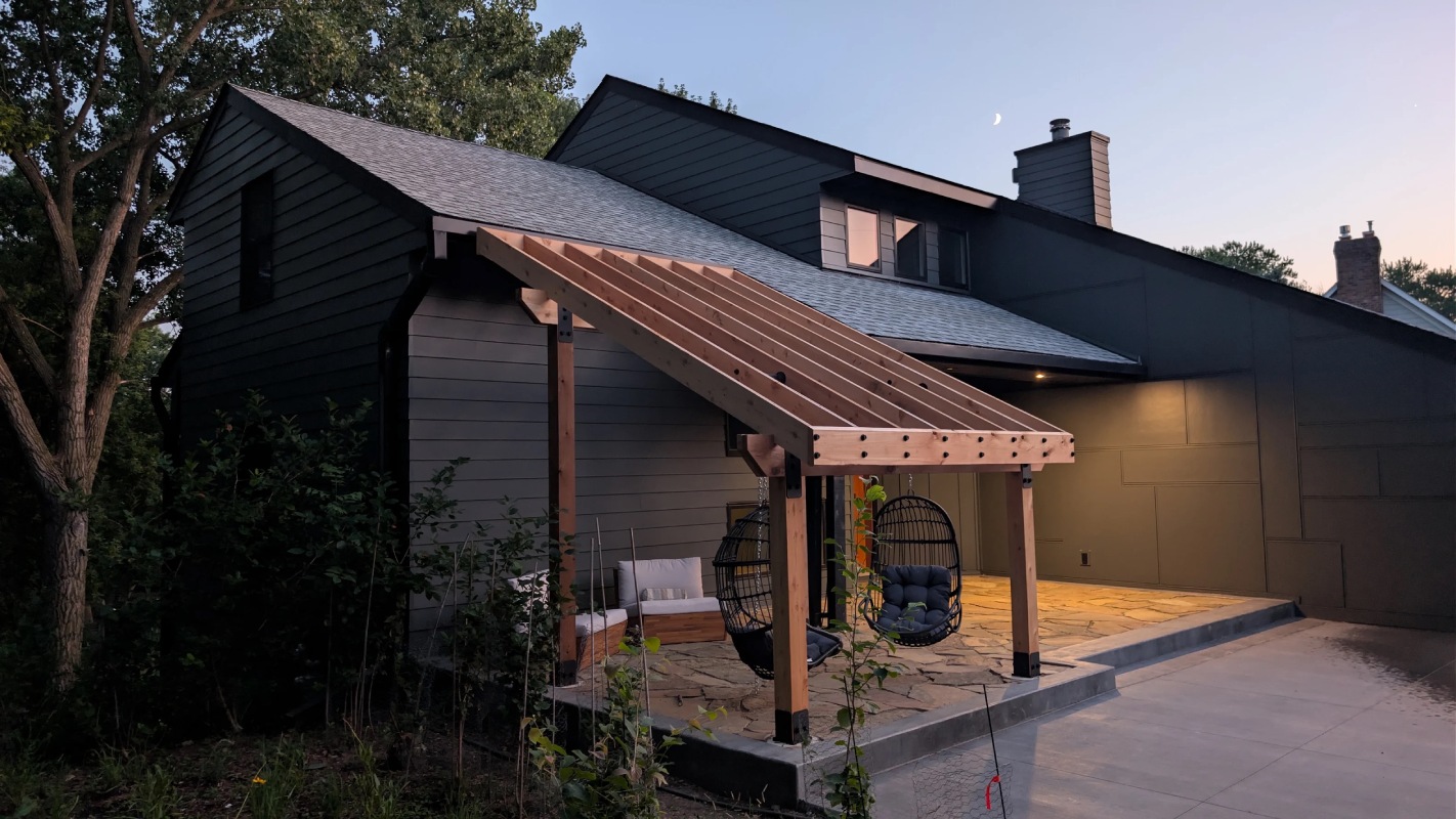 Cedar pergola with a sloped roof frame attached to a dark-sided house, overlooking a stone patio at dusk.