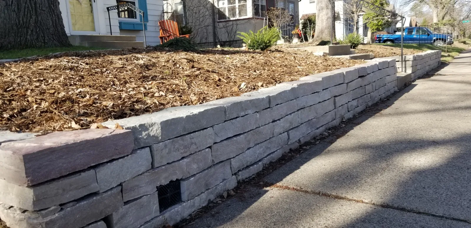 Low stacked natural stone retaining wall with mulched planting bed and drain opening beside a sidewalk in Eagan, MN