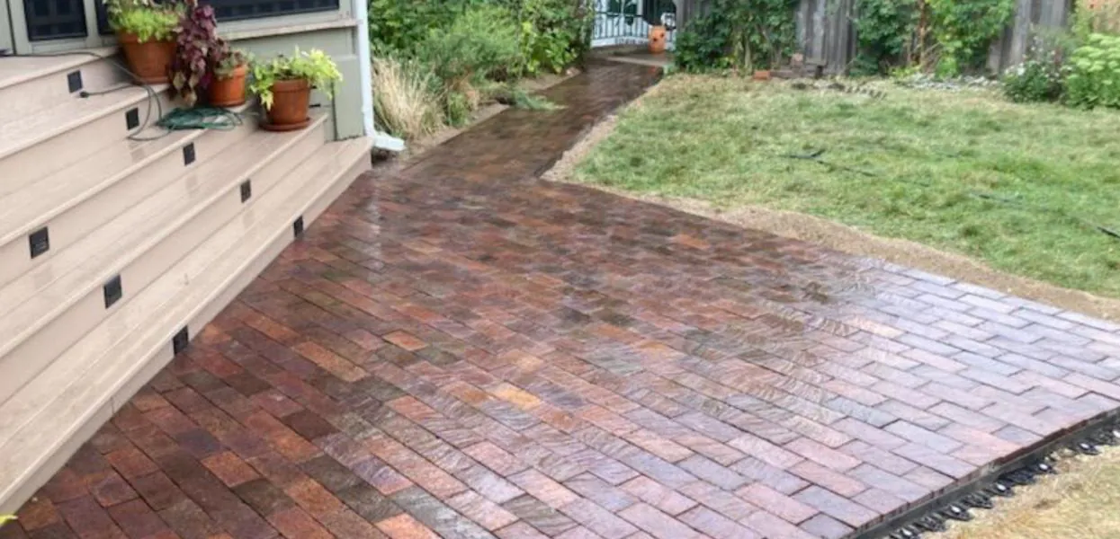 Red brick paver walkway curving past wood deck steps and potted plants in a St. Paul, MN backyard